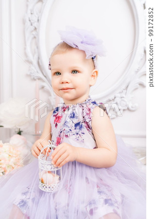 Baby girl elegant dress. A one-year-old girl in a puffy dress and a cute bow poses against the backdrop of a bright room with a dressing table and flowers. Baby girl elegant dress. A one-year-old girl in a puffy dress and a cute bow poses against the backdrop of a bright room with a dressing table and flowers. 99172852