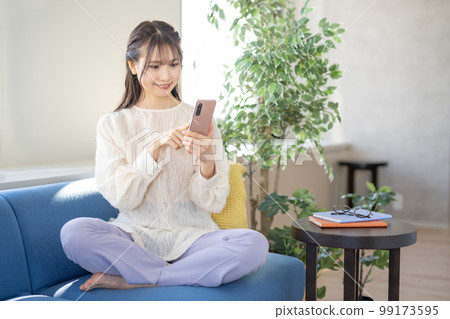 A young woman sitting cross-legged on the sofa and operating a smartphone 99173595