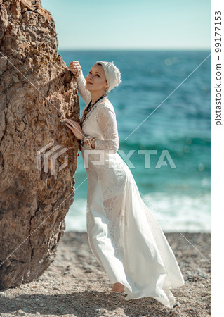 Woman white dress sea stones rocks.Middle-aged woman looks good with blond hair, boho style in a white long dress on beach jewelry around her neck and arms. 99177153