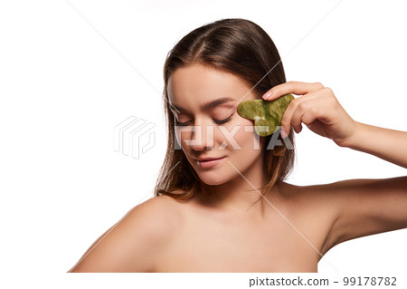 Young beautiful girl massaging face with Gua sha over white studio background. Circulation and lifting effect. Toned and smooth skin  99178782