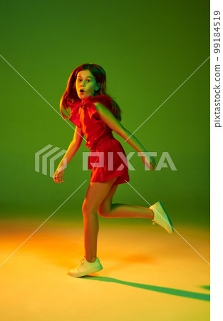 Active playful kid. Little girl, child in red clothes posing over gradient green yellow studio background in neon light. Concept of childhood and emotions 99184519