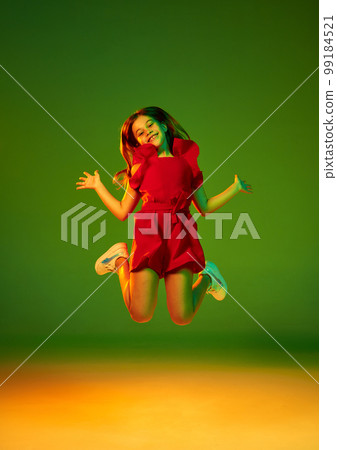 Little girl, child in red clothes posing, cheerfully jumping over gradient green yellow studio background in neon light. Concept of childhood and emotions 99184521