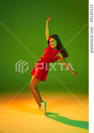Little girl, child in red clothes posing, dancing over gradient green yellow studio background in neon light. Concept of childhood and emotions 99184523