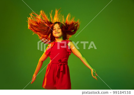 Little girl, child in red outfit posing with happy positive face over green studio background in neon light. Concept of childhood and emotions 99184538