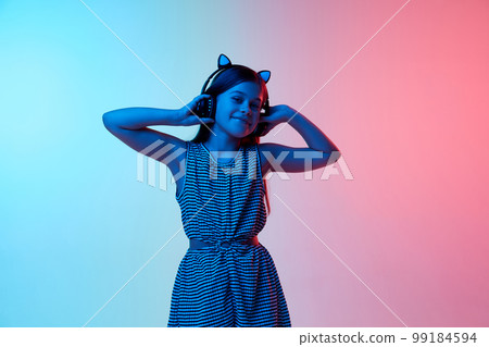 Little beautiful girl, child listening to music in headphones over gradient blue pink studio background in neon light. Concept of emotions and childhood 99184594