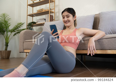 young woman exercising at home happily sitting and resting playing on the phone 99184824