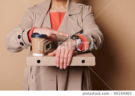 It's lunch time. Close-up woman holding takeaway pizza and coffee in paper mug, points her index finger at a wristwatch It's lunch time. Close-up woman holding takeaway pizza and coffee in paper mug, points her index finger at a wristwatch 99184865