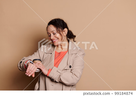 Beautiful woman checking time on her wristwatch, happy to be punctual for a meeting, isolated on beige background Beautiful woman checking time on her wristwatch, happy to be punctual for a meeting, isolated on beige background 99184874