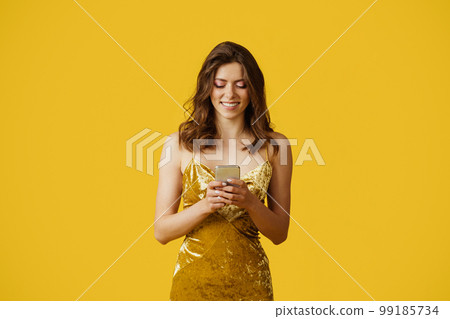 Cheerful lady in dress typing on smartphone, reading message, standing on yellow background, studio shot 99185734