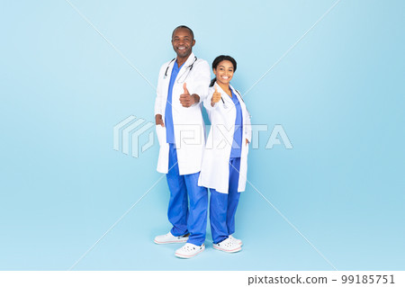 Cheerful black female and male therapists in white coats showing thumbs up, blue studio background, full length Cheerful black female and male therapists in white coats showing thumbs up, blue studio background, full length 99185751