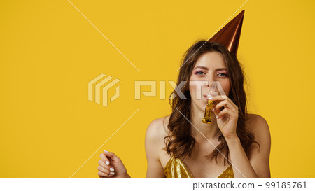Pretty caucasian woman in birthday cap blowing party horn, celebrating her special day, yellow background, free space Pretty caucasian woman in birthday cap blowing party horn, celebrating her special day, yellow background, free space 99185761