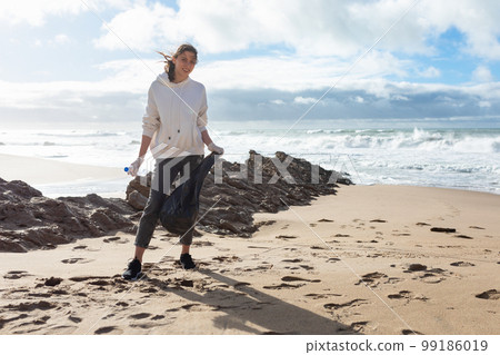 World environment day. Female volunteer standing on beach with bag, picking up garbage and cleaning the coastal zone World environment day. Female volunteer standing on beach with bag, picking up garbage and cleaning the coastal zone 99186019