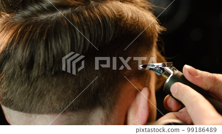 close up Shot of a handsome barber giving a haircut to his client using trimmer. Hairdresser service in a modern barbershop in a dark key lightning with warm light back view close up Shot of a handsome barber giving a haircut to his client using trimmer. Hairdresser service in a modern barbershop in a dark key lightning with warm light back view 99186489