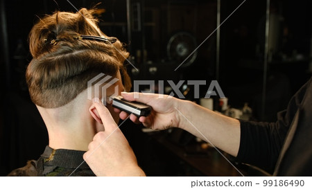men's hairstyling and haircutting with barber shaving machine, hair cut for male. Hairdresser service with shaver in a modern barbershop in a dark key lightning with warm light back view men's hairstyling and haircutting with barber shaving machine, hair cut for male. Hairdresser service with shaver in a modern barbershop in a dark key lightning with warm light back view 99186490