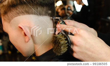 Shot of a handsome barber giving a haircut to his client using trimmer. Hairdresser service in a modern barbershop in a dark key lightning with warm light side view 99186505
