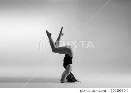 Young slim female in black bodysuit makes yoga stretching exercise on white studio wall background. Young slim female in black bodysuit makes yoga stretching exercise on white studio wall background. 99186804