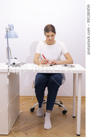 Nail beautician makes notes in notebook with pen, sitting at workstation, table in nail beauty treatment salon. Professional tools are on white table. Manicurist's routine. Vertical 99186956