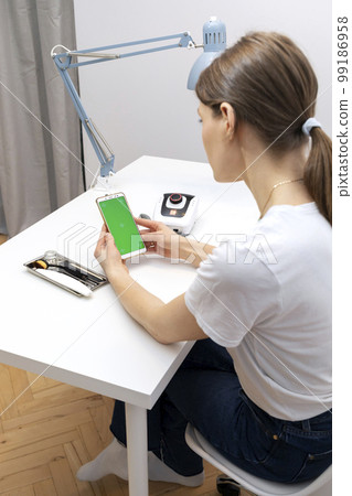 Manicure, pedicure professional holds smartphone with green chroma key screen, sitting at workstation in nail beauty treatment salon.Professional tools are on white table.Studying manicurist. Vertical 99186958