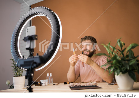 Portrait of happy man beauty blogger sitting in room at table and speaking recommending foundation or decorative cosmetic looking at camera. Blogging and vlog concept Portrait of happy man beauty blogger sitting in room at table and speaking recommending foundation or decorative cosmetic looking at camera. Blogging and vlog concept 99187139