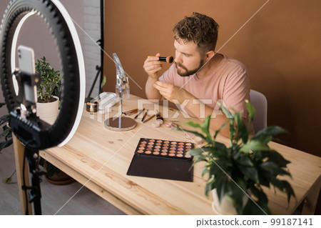 Man talking on cosmetics holding a makeup tools while recording his video. Guy making video for his blog on cosmetic product Man talking on cosmetics holding a makeup tools while recording his video. Guy making video for his blog on cosmetic product 99187141
