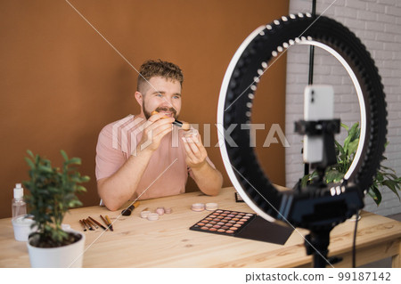 Male beauty blogger doing his make up and recording a video lesson his with mobile phone - vlog, diversity and video blog concept Male beauty blogger doing his make up and recording a video lesson his with mobile phone - vlog, diversity and video blog concept 99187142