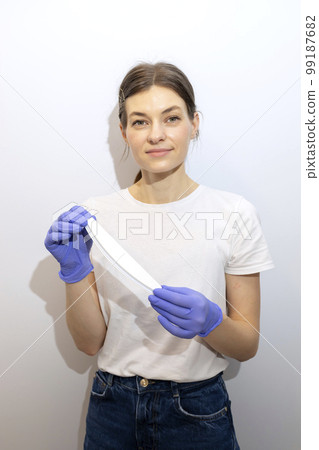 Caucasian manicurist, nail technician holds file in hands, wearing gloves. White smiling manicure master stands near gray wall in spa salon. Soft, safe beauty nail care, treatment. Vertical plane 99187682