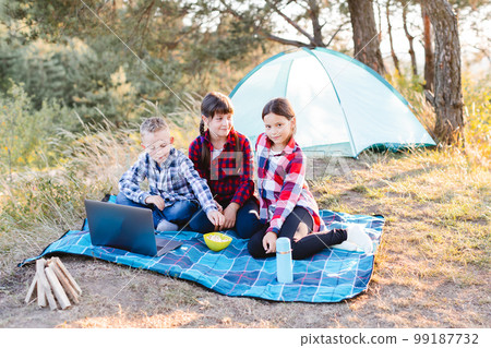 Children looking show on laptop in the forest. Boys and girls are resting in a tent camp. Children sit on the plaid on the grass in summer. 99187732