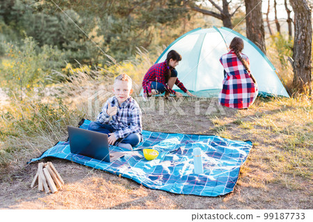 Lets go camping. Family camping. Reach destination place. Two girls pitch tent and one boy watching show on the laptop, sitting on the plaid. Wanderlust discovery. Hiking outdoor adventure. 99187733