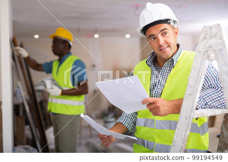 Builder in yellow vest checks the drawings in renovated room 99194549