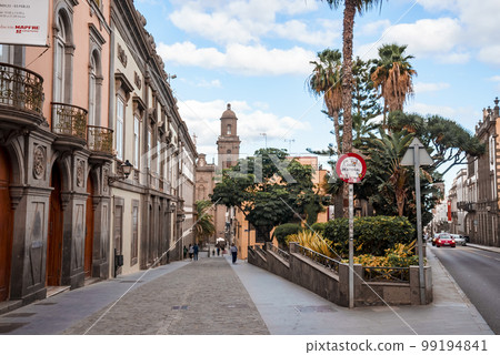 Diminishing perspective of narrow street between historic buildings and trees leading towards famous Cathedral of Santa Ana at Las Palmas. Cloudy day in Gran Canaria Islands at Spain 99194841