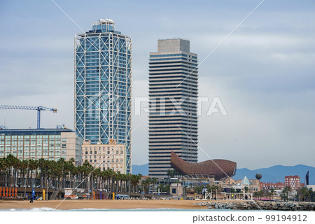 View of Frank Gehry's Golden Fish Sculpture with Mapfre Tower and Hotel Arts at Port Olympic with cloudy sky in the background at Catalonia in Barcelona, Spain View of Frank Gehry's Golden Fish Sculpture with Mapfre Tower and Hotel Arts at Port Olympic with cloudy sky in the background at Catalonia in Barcelona, Spain 99194912