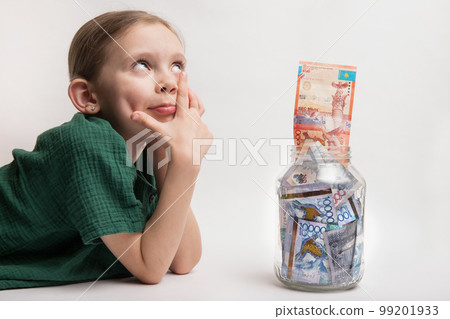 Happy girl with a large transparent jar with Kazakhstani tenge money. the child thought about new toys and gadgets Happy girl with a large transparent jar with Kazakhstani tenge money. the child thought about new toys and gadgets 99201933