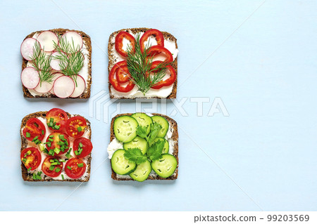 Variety of sandwiches for breakfast - slice of whole grain dark bread, pepper, cream cheese, cucumbers, radishes, cherry tomatoes, garnished with dill, green onions on blue table Top view Flat lay. Variety of sandwiches for breakfast - slice of whole grain dark bread, pepper, cream cheese, cucumbers, radishes, cherry tomatoes, garnished with dill, green onions on blue table Top view Flat lay. 99203569