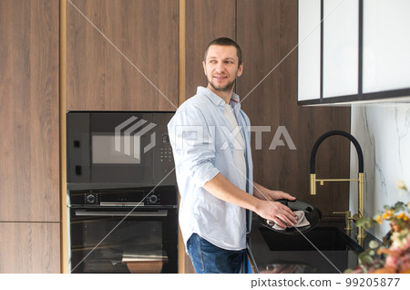 A man in a shirt wipes dishes with a towel standing in the kitchen. Household chores A man in a shirt wipes dishes with a towel standing in the kitchen. Household chores 99205877