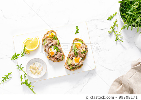 Tuna toast. Open sandwiches with whole grain bread, canned tuna, boiled egg, avocado and arugula. Top view Tuna toast. Open sandwiches with whole grain bread, canned tuna, boiled egg, avocado and arugula. Top view 99208171