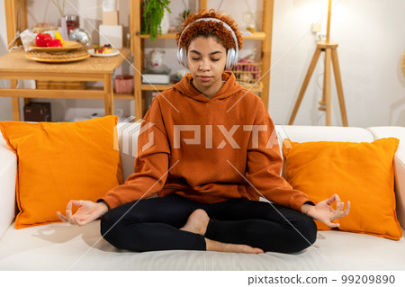 Yoga mindfulness meditation. Young healthy african girl practicing yoga at home. Woman sitting in lotus pose on couch meditating smiling relaxing indoor. Girl doing breathing practice. Yoga at home Yoga mindfulness meditation. Young healthy african girl practicing yoga at home. Woman sitting in lotus pose on couch meditating smiling relaxing indoor. Girl doing breathing practice. Yoga at home 99209890
