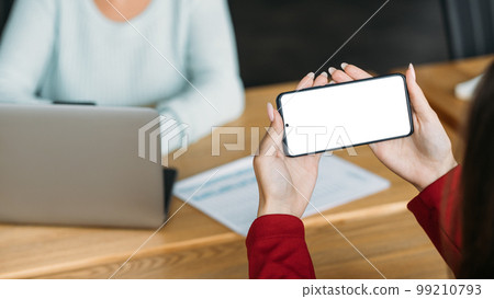 Online presentation. Mobile mockup. Office routine. Female hands holding smartphone with blank screen sitting with colleague in light room interior. 99210793