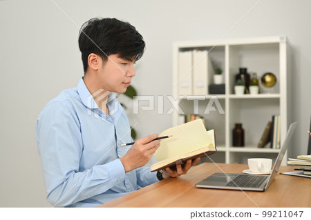 Young businessman looking at laptop screen and making important notes on notepad 99211047