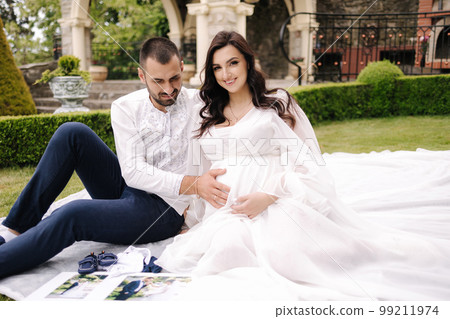 Beautiful couple sitting on a plaid on the grass. Elegant pregnant woman with husband waiting for baby. Future parents 99211974