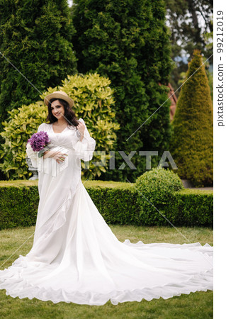 Elegant pregnant woman in beautiful white dress walking outdoors. Future mom hold wicker basket with flowers 99212019