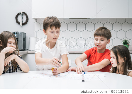 Happy little school friends play board game on paper map with dice and chips at kitchen table. Entertaining table game. 99213921