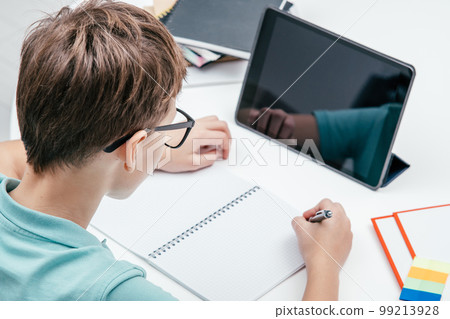 Top view of preteen boy sitting at desk in front of tablet, having online lesson, writing on notebook with left hand. 99213928