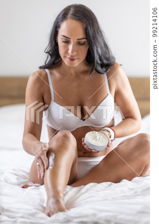 Beautiful woman applying body-hydrating cream on her leg in bedroom 99214106