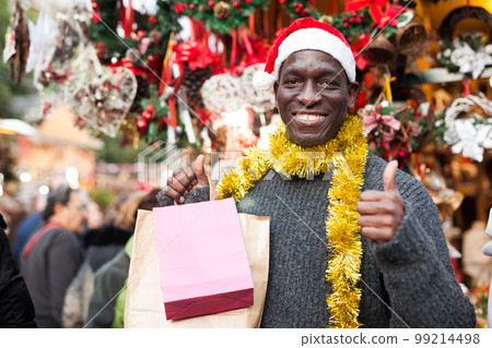 Smiling man in Santa hat with shopping bags on Christmas fair Smiling man in Santa hat with shopping bags on Christmas fair 99214498