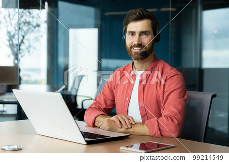 Portrait of mature businessman in red shirt inside office, man with beard and video call headset smiling and looking at camera, online customer support worker with laptop. Portrait of mature businessman in red shirt inside office, man with beard and video call headset smiling and looking at camera, online customer support worker with laptop. 99214549