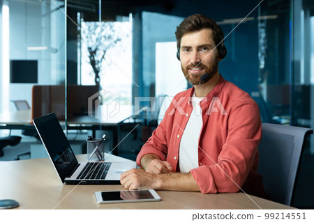 Portrait of mature businessman in red shirt inside office, man with beard and video call headset smiling and looking at camera, online customer support worker with laptop. Portrait of mature businessman in red shirt inside office, man with beard and video call headset smiling and looking at camera, online customer support worker with laptop. 99214551