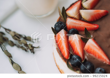 Bird's milk cake with chocolate decorated with strawberries and grapes on white table with glass of milk 99215024