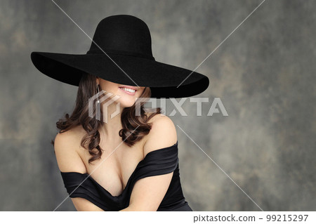 Beautiful woman brunette wearing black dress and wide broad brim hat posing on gray background 99215297