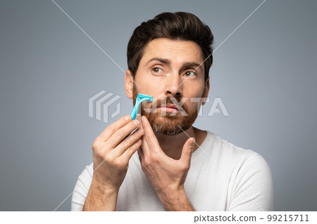 Handsome bearded middle aged man in t-shirt shaving his beard while standing over grey studio background 99215711
