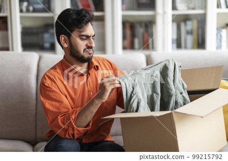 Disappointed middle eastern man customer opening parcel at home Disappointed middle eastern man customer opening parcel at home 99215792